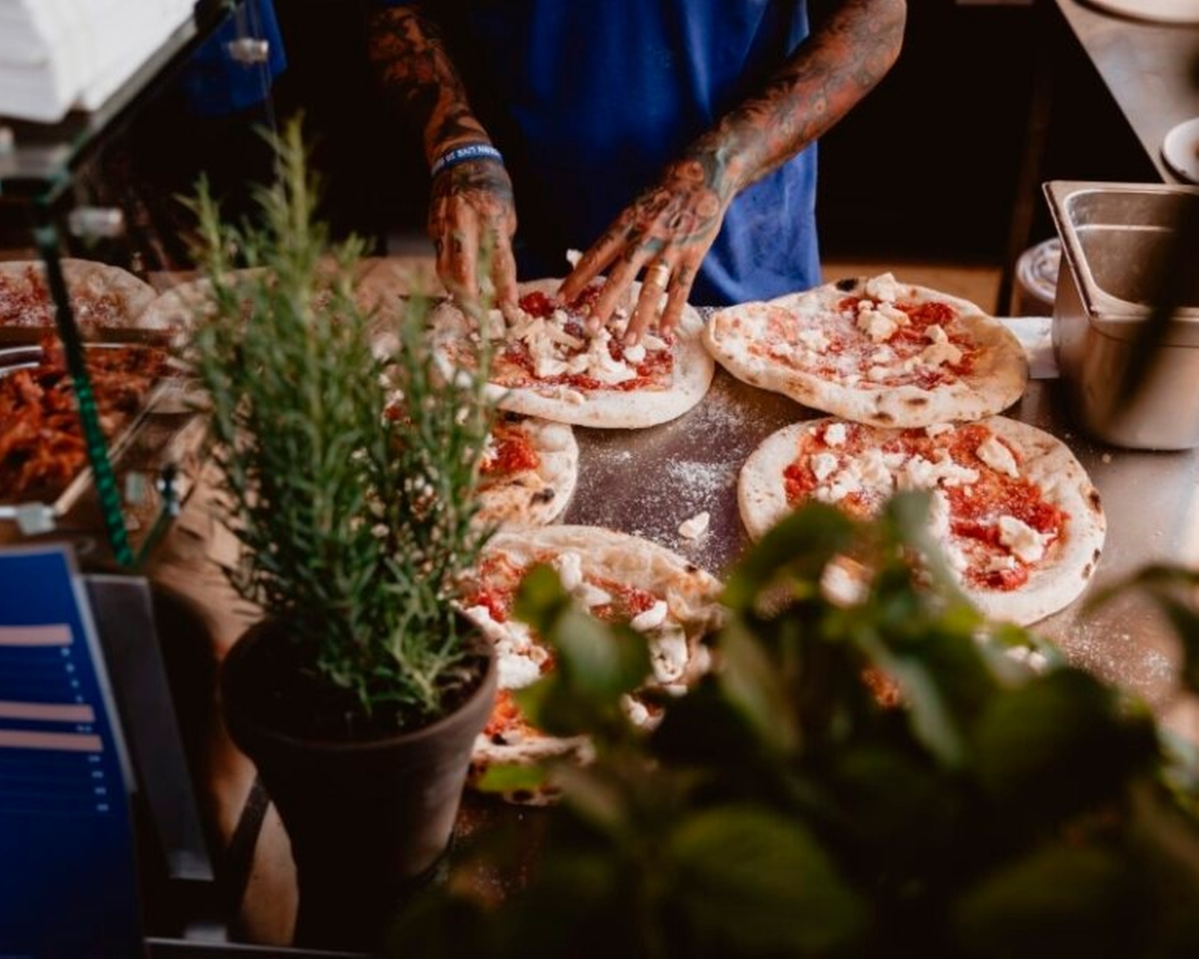 Koch bereitet Pizzen in einer Küche mit frischen Zutaten und Kräutern im Vordergrund zu