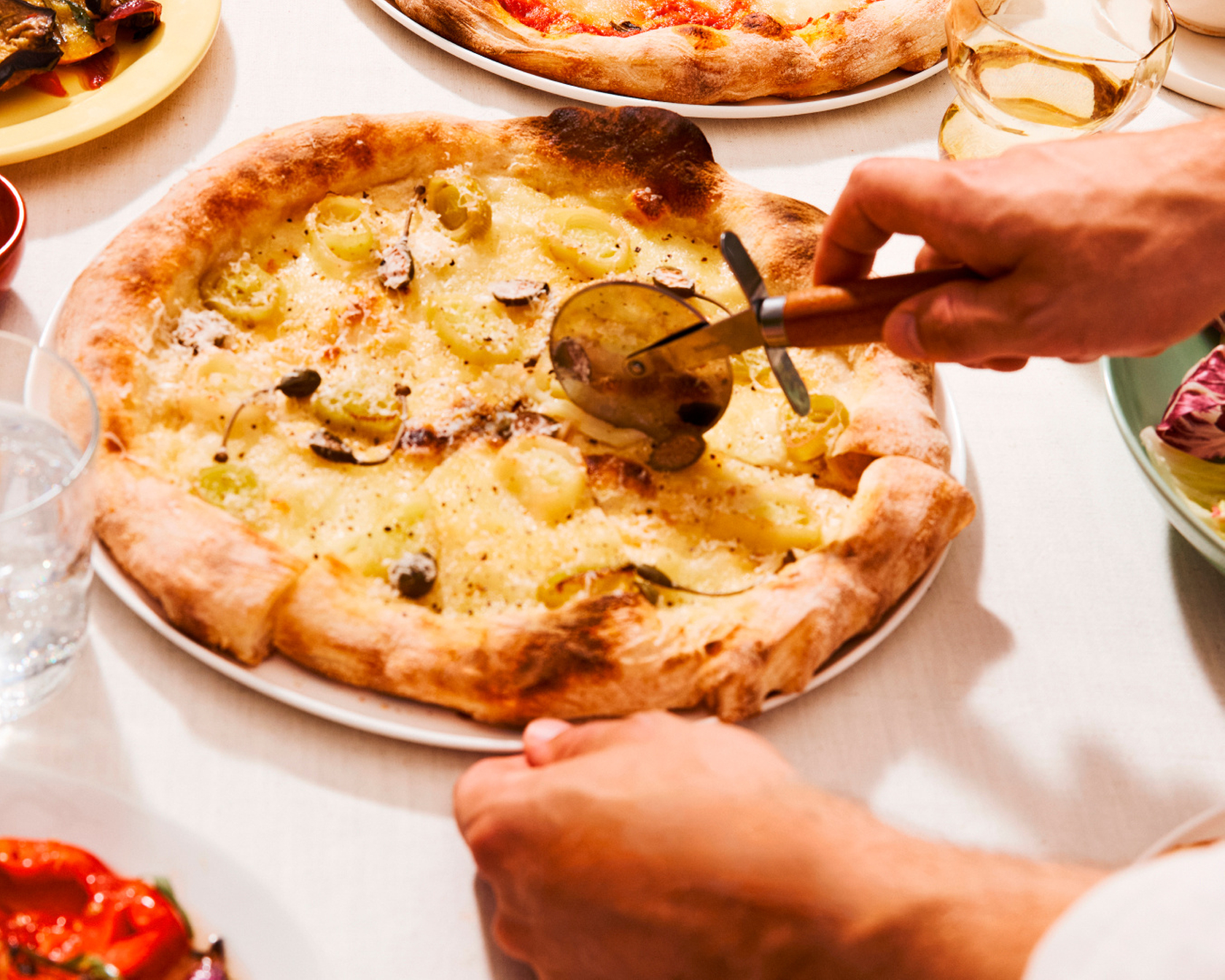 Frisch gebackene Pizza mit Kartoffeln und Kapern, die am Tisch mit einem Pizzaschneider geschnitten wird.