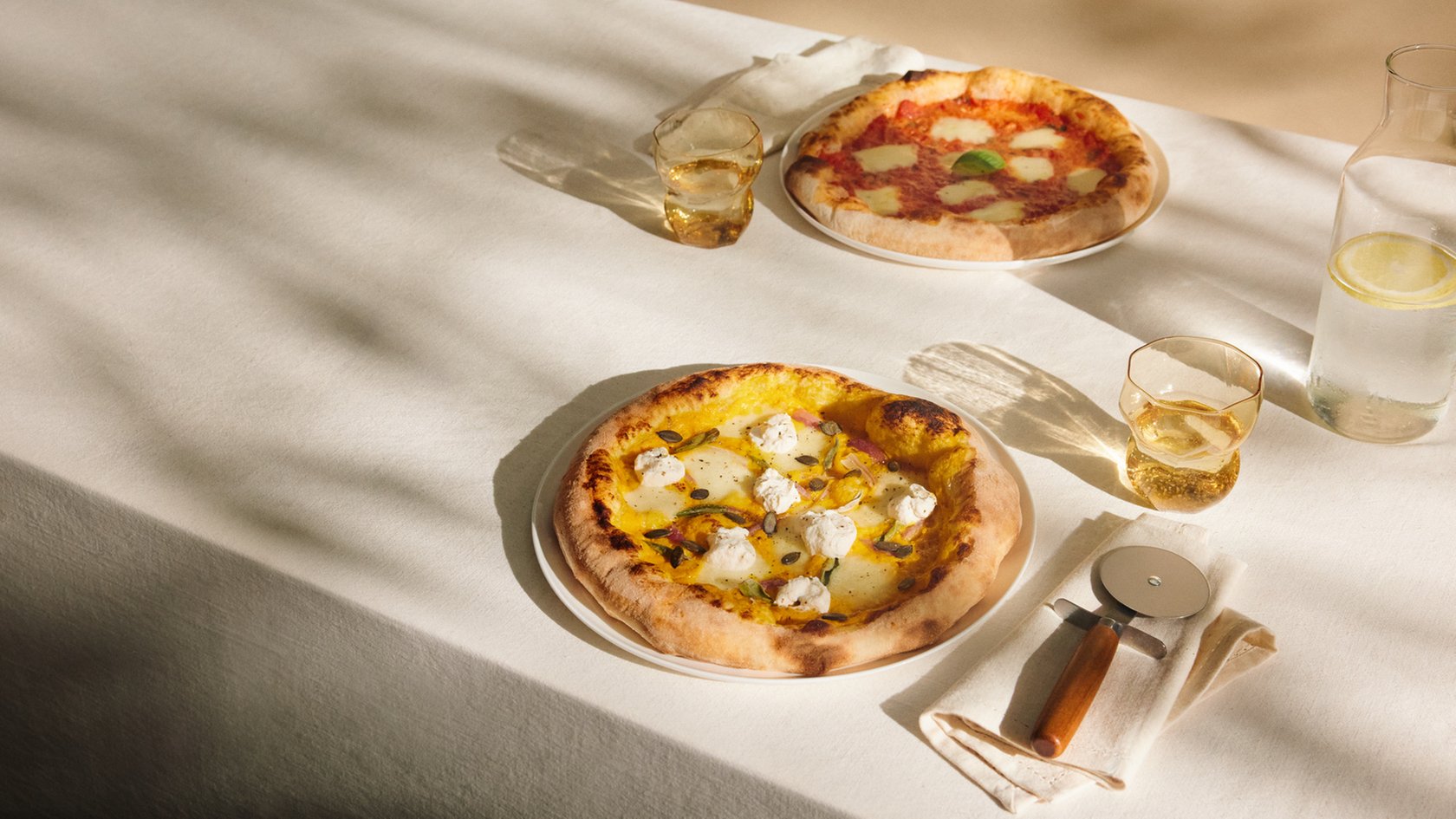 Zwei Pizzen auf einem Tisch mit Getränken und Pizzaschneider in natürlichem Licht.