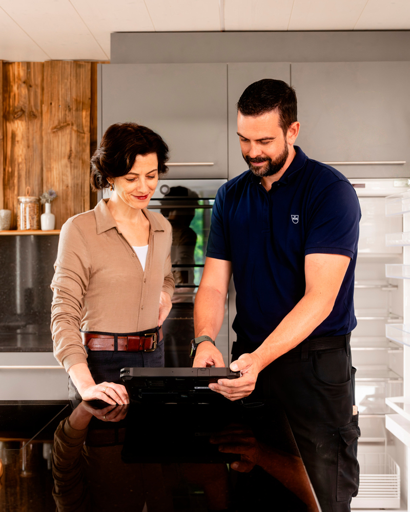 Consultation dans une cuisine moderne avec un réfrigérateur ouvert et un conseiller qui montre une tablette.