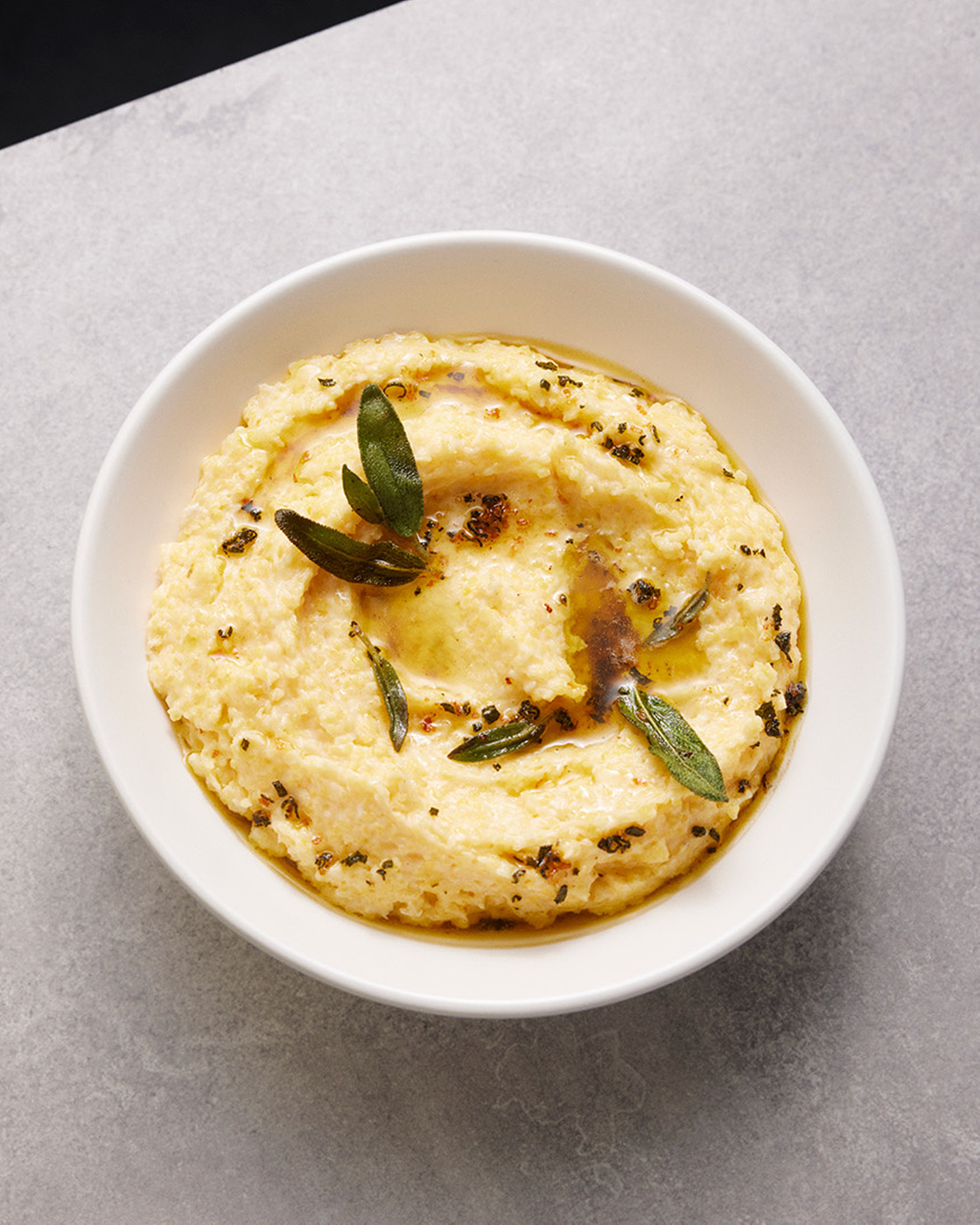 Cremige Polenta mit Kräutern in einer weißen Schale auf grauem Hintergrund.