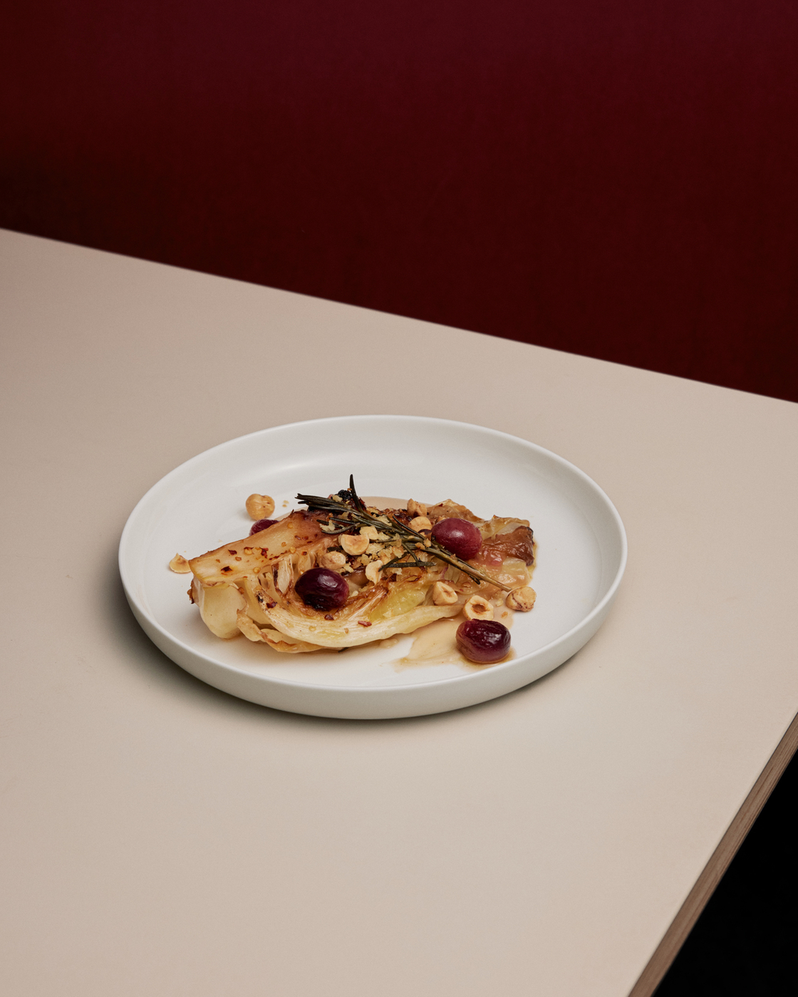 Roasted chicory with grapes and nuts on a white plate, served on a table against a dark red background.