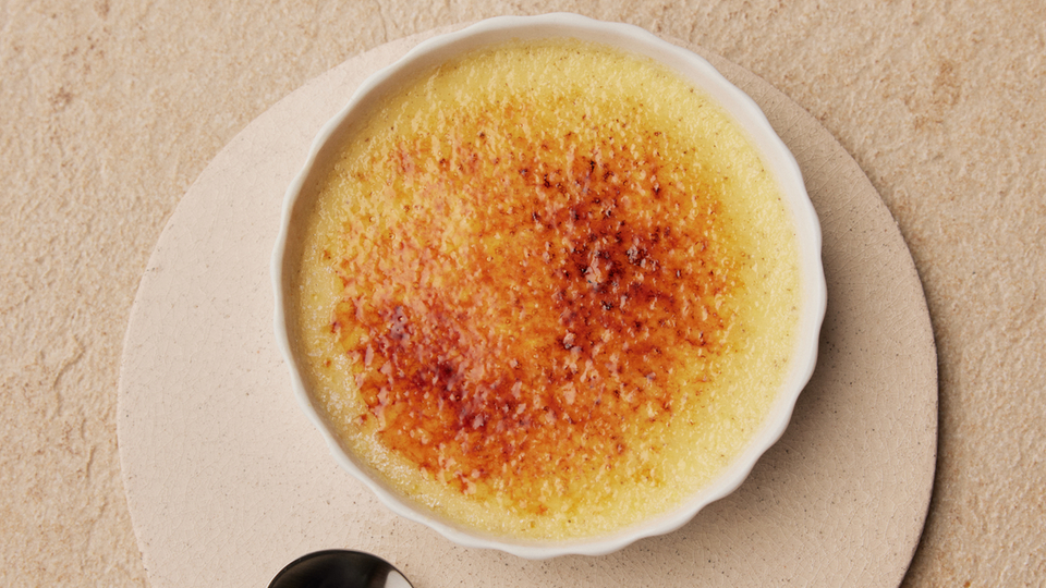 Crème Brûlée in einer weißen Schale mit karamellisierter Oberfläche und einem Löffel auf einem Untersetzer.