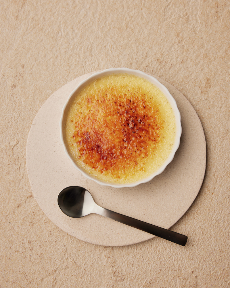 Crème brûlée servie dans un bol blanc avec une surface caramélisée et une cuillère sur un dessous de plat.