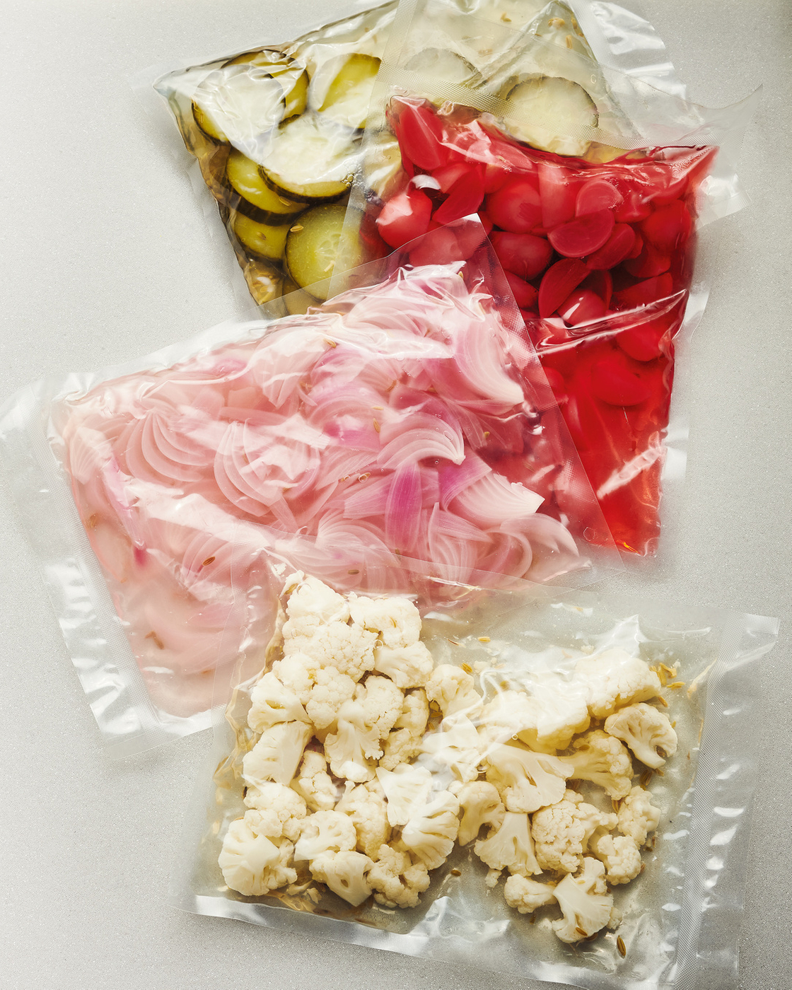 Vacuum-packed vegetables such as cauliflower, onions and pickled cucumbers on a light-colored surface.