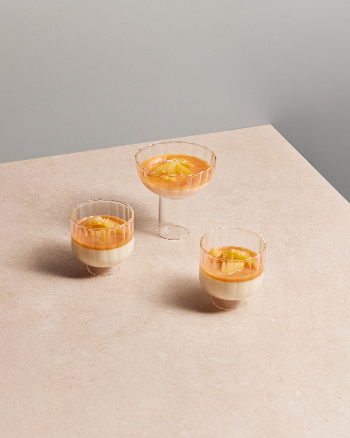 Drei Dessertgläser mit Creme und orangefarbenem Fruchtspiegel auf hellem Tisch in minimalistischer Umgebung.