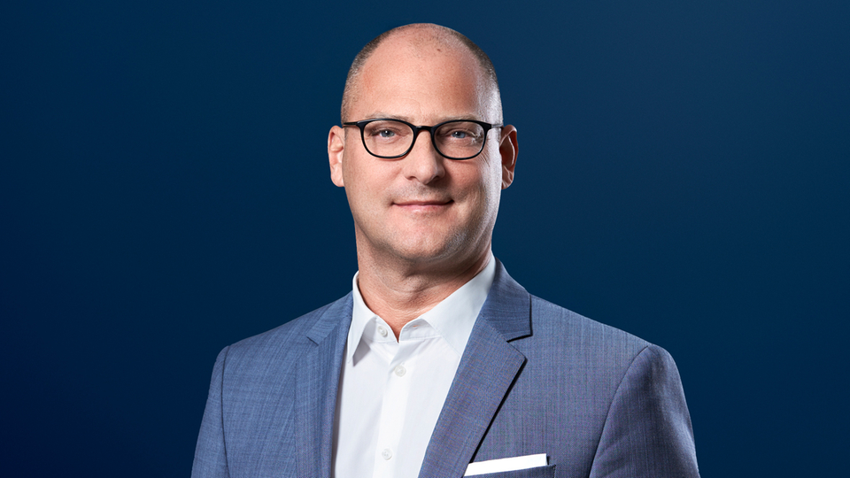 Portrait of a man wearing glasses in a light blue suit against a dark blue background